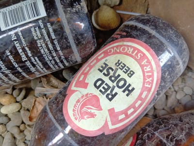 This image shows three empty beer bottles scattered on the ground, which appears to be a mix of small stones and dirt. One bottle, prominently featured in the foreground, is a "Red Horse Beer" bottle with its distinctive red and white label. The label is partially obscured by dust and dirt. The other two bottles are also partially visible, and appear to be similar dark glass beer bottles. The overall scene suggests a discarded collection of empty beverage containers in an outdoor setting. Given the "San Miguel Brewery" and "Philippines" text visible on one of the bottles, it is possible this scene is located in the Philippines. However, no specific location in the city of Samal, Philippines can be confirmed from the visual evidence alone.