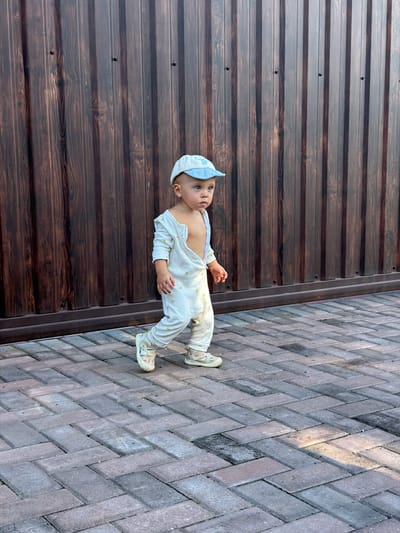 A baby is lying on a patterned tiled path next to a dark wooden fence. The baby is wearing a white onesie with a yellow fruit pattern, a white cap, and light-colored shoes. The baby's eyes are open, and they are looking upwards. The scene appears to be outdoors, with natural light suggesting it is daytime. The wooden fence has horizontal planks, and the tiled path is laid in a herringbone pattern. The location context is Борисьево Днп, Russia.