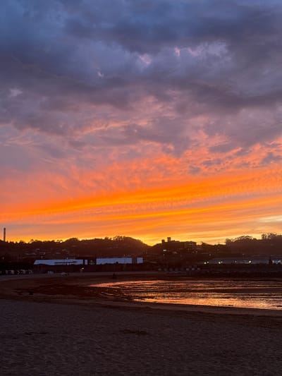 The image depicts a sunset over the city of Gijón, Spain, specifically the area near El Musel port. In the foreground, a sandy beach is visible, with the dark sand contrasting with the reflective water in the middle ground. The water appears calm, with ripples reflecting the vibrant colors of the sky. Beyond the water, a strip of land with buildings and industrial structures lines the coast. A tall, narrow smokestack is prominent on the left side of this strip. In the background, a tree-covered hillside rises, dotted with some buildings. The sky is the dominant feature, ablaze with intense orange and yellow hues from the setting sun, transitioning into purple and gray clouds higher up. The overall scene captures a dramatic and colorful end to the day.