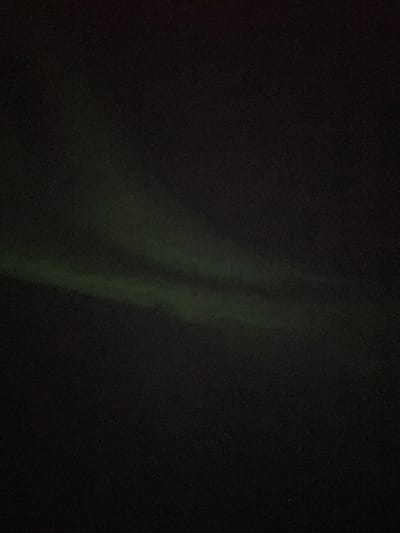 The image shows a night sky, primarily dark, with a faint display of the Aurora Borealis. The aurora appears as a vertical, soft-glowing green band, slightly diffused and irregular in shape. The rest of the sky is mostly black, with subtle variations in darkness hinting at other atmospheric phenomena. A few faint stars may be visible. The overall composition evokes a sense of mystery and natural beauty.