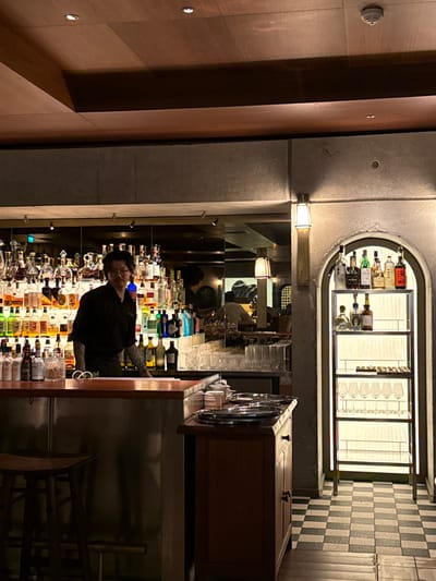 The image portrays the interior of Le Bar, located within the InterContinental Bordeaux - Le Grand Hôtel at 2-5 Place de la Comédie, 33000 Bordeaux, France. A male bartender, with dark hair and visible tattoos on his forearms, stands behind a stainless steel and wooden bar counter in the left-center of the frame, facing the viewer with a slight smile. He is wearing a black short-sleeved shirt.

Behind the bartender, an extensive array of liquor bottles is displayed on multiple tiered, brightly backlit shelves, serving as a prominent feature of the bar. Various bar implements, including glassware and a container of nuts, are visible on the bar surface. To the immediate right of the bar, a wooden service cabinet with two closed doors holds several stacked silver serving trays. A dark wooden bar stool with a round seat is present in the lower-left foreground.

The right side of the scene features a textured concrete-like wall with a vertical wall-mounted sconce emitting a warm light. Below this, an arched, internally illuminated niche displays additional liquor bottles on its top shelves and a row of clear wine glasses on its bottom shelf. The floor in this section exhibits a black and white checkerboard tile pattern. The ceiling above is a multi-tiered wooden structure with recessed lighting. A mirror behind the main bar reflects a partial view of another individual in the background and a rectangular-shaded lamp.