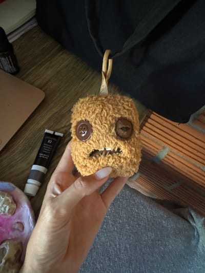 A handmade, fuzzy, orange creature with two buttons for eyes and a sewn mouth is held by a person's hand. The creature has a small loop attached to its head. In the background, a dark fabric bag, a wooden surface, a tube of cosmetic cream, and a brick wall are visible. There is no indication of the image being taken in Baan Nok Sai, Thailand.