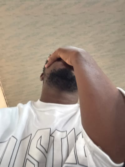 The scene is located in Osu, Nigeria, and features an adult male captured from a low-angle, upper-chest perspective, looking upwards. He has a dark skin tone, short dark hair, and a trimmed dark beard. He is dressed in a white, long-sleeved crew-neck shirt with the partial word "JUST" outlined in dark characters on the front. His right hand is raised, covering his forehead and upper face. The background is a light-colored, subtly patterned ceiling or upper wall.