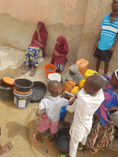 **People:** A group of children and adults, approximately seven individuals, are depicted. Some wear traditional clothing, including headscarves and patterned fabrics. One individual wears a blue and white striped shirt.
**Subjects:** The focus is on the process of collecting water.
**Objects:** Several containers are present, including buckets, plastic jerrycans (yellow and blue), and metal water vessels.
**Actions:** The individuals are gathered around a water source, filling containers. Some appear to be pouring water.
**Interactions:** People seem to be working together to collect water.
**Environment:** The scene appears to be a communal water point in Dutsinma A, Nigeria. The ground is wet and muddy, indicating recent water spillage. The backdrop consists of a simple structure with a partially painted wall.