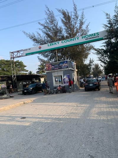 The scene depicts the main entrance to Lekky County Homes, located in Ikota, Lagos, Nigeria. A prominent metal archway spans a paved road, displaying a white and green sign with "LEKKY COUNTY HOMES RESIDENTS" and "CREATED BY" alongside a red and white logo. To the right of the entrance, a small, grey concrete security booth with windows and various advertisements affixed to its exterior is visible. Approximately seven to ten adult males are gathered around this booth; several are seated on the ground or a low ledge, while others stand nearby.
On the left side of the entrance, behind a parked dark-colored SUV, several small commercial structures or stalls with corrugated metal roofs are present. A black sedan is visible further down the road to the right, positioned as if entering or exiting the estate. The sky is clear blue, and tall, slender evergreen trees line both sides of the road and extend into the background. Overhead power lines are visible against the sky. The foreground consists of a grey, textured paved road surface, with concrete curbs defining its edges. Natural sunlight illuminates the scene, casting distinct shadows.