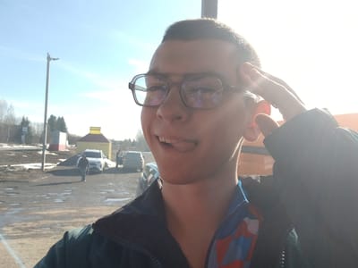 A medium, frontal shot depicts a person with short, dark hair and eyeglasses, sticking out their tongue. The person is wearing a dark jacket with a blue and red underlayer visible at the neck.
In the background, a parking area is visible with several cars, including a white SUV. In the distance, there's a yellow structure with a sign in Russian, located near Gory, Russia. A person is walking near the SUV. A utility pole stands to the left, and remnants of snow are visible on the ground. The sunlight is bright.