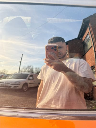 A male subject is captured in a reflection, holding a smartphone in his right hand to take a selfie. He wears a white short-sleeved t-shirt and has short dark hair with faded sides. His face is partially obscured by the rose-gold/orange smartphone, which features a pop socket. Visible on his right arm are irregular patches of lighter skin, consistent with vitiligo. The reflection is cast upon a dirty glass surface, possibly a vehicle window, given the orange panel visible at the bottom.
The background, visible through and beyond the reflection, depicts an outdoor scene in Bretton, United Kingdom, under a clear, bright blue sky with sparse, wispy clouds. To the right, a red-brick building with dark-framed windows is partially visible. To the left, a white panel van, possibly a Vauxhall Combo, is parked on an asphalt or gravel surface. Behind the van, a wooden utility pole with multiple crossbars and insulators stands. Leafless trees are visible in the distant background.