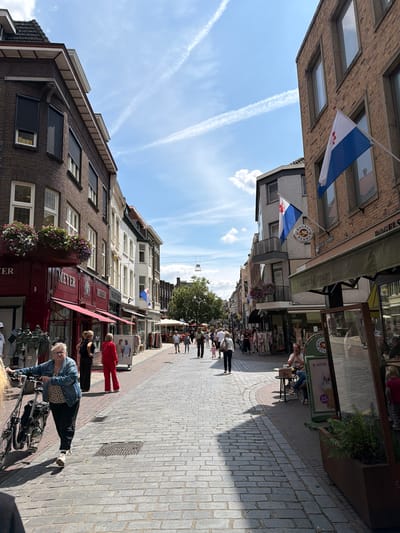 The image depicts a street scene in the city of Roermond, Netherlands. The street is paved with cobblestones and lined with buildings. On the left, a dark brick building with large windows and awnings houses a store called "MEYER". Flower boxes filled with colorful flowers are attached to the building. Several people are walking along the street.

Further down the street, on the right side, a brick building has a flag flying from it. Next to it, a store called "BAGELS & BEANS" is visible, with awnings extending outwards. More people are seen walking and sitting at tables outside the store. The sky is blue with scattered white clouds and contrails from airplanes. The overall scene suggests a busy urban environment on a sunny day.