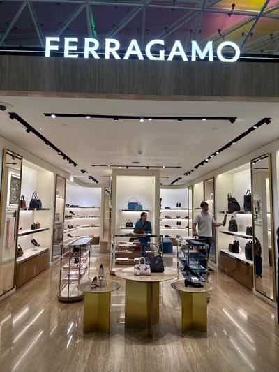 The image shows the interior of a Ferragamo store, likely located in a shopping mall. The store is well-lit with bright overhead lighting and display lights illuminating the merchandise. The floor is made of polished marble tiles with a reflective surface.
In the foreground, there are two round display tables with gold-colored bases. The left table holds a light pink handbag, and the right table displays a pair of beige high-heeled shoes. A narrow display rack with multiple shelves containing handbags is visible behind the right table.
Further into the store, there are numerous shelves stocked with shoes and handbags. A woman in a teal blazer is standing behind a counter on the left side of the image, and a man in a light grey polo shirt is standing in the middle of the store, holding a black shoe.
The store's name, "FERRAGAMO," is prominently displayed in large white illuminated letters above the entrance. The background shows the colorful, iridescent ceiling of what appears to be a mall.
The location is the city of Nong Prue Subdistrict, Thailand.