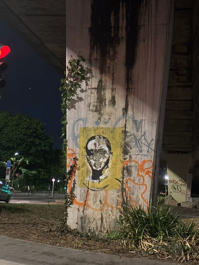 Here is a description of the image:
The photograph shows a concrete pillar supporting an overpass, likely in an urban environment. The pillar features extensive graffiti, including an abstract portrait painted within a yellow square and orange looping lines. The base of the pillar is flanked by vegetation: grass and weeds on one side and climbing ivy on the other. Beyond the pillar, one can see a street at night, with streetlights, traffic lights, and trees lining the roadside. The sky is dark, indicating it is nighttime. Based on some of the graffiti in the image and its style, it may depict a location in the city of Manchester, United Kingdom, although the image does not provide enough clear information to confirm this with absolute certainty.