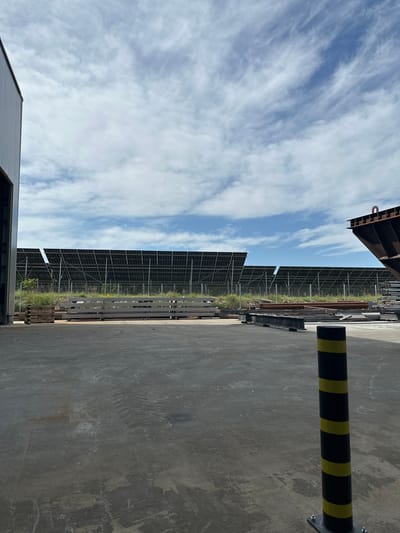 The image shows a ground-level perspective of an industrial site, possibly in Vedrare, Bulgaria, given the prompt. A row of solar panels stretches into the distance under a cloudy sky. In the foreground, there's a gray concrete wall or surface and a black pole with yellow stripes attached to the surface with metal brackets. Metal beams and other industrial materials rest on the ground between the solar panels and the foreground. A section of a building roofline is visible at the bottom left. At the top left, a dark-colored industrial structure is partially visible. There are no people visible in the image. The environment suggests an outdoor industrial area.