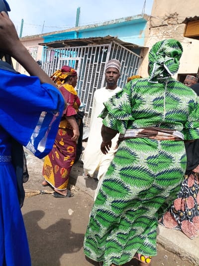 The image depicts an outdoor street scene in Gama, Nigeria, with multiple individuals. In the foreground, partially visible on the left, is a person wearing a blue garment with white trim. On the right, a woman in a green and dark blue leaf-patterned dress is seen from behind, with a brown fabric sash tied around her waist. In the mid-ground, a bearded man wearing a light-colored robe and a patterned cap (kufi) looks directly at the viewer. To his left, a woman in a maroon, yellow, and gold patterned dress and a matching headwrap is partially obscured, facing away from the camera. Another individual with a patterned cap is partially visible in the background to the right. The ground is unpaved dirt, showing minor debris, including a bundle of sticks near the left mid-ground woman's feet and a small light-colored stone in the center. In the background, a light blue building with a corrugated metal overhang and a white metal security gate is visible. Two green poles extend from the building's roof, and a faded sign with partial text is present on its wall. The sky above is light blue.