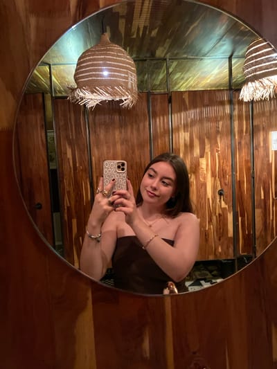 A medium shot shows a woman, wearing a brown top, reflected in a round mirror. She is holding a cell phone with a patterned case and is taking a selfie. She has brown hair and is wearing jewelry. The background behind the mirror reflection is a wooden wall with horizontal planks. In the upper left quadrant of the mirror's reflection, a hanging wicker light fixture is visible. The photo appears to have been taken indoors, and the location is likely in Oaxaca City, Mexico, given the prompt.