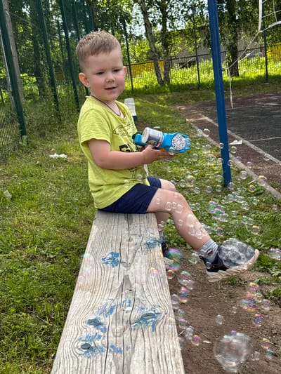 A young boy with short, light brown hair is sitting on a weathered wooden bench in Zav'yalovo, Russia. He is wearing a neon yellow t-shirt with a graphic print and dark blue shorts. The boy is holding a blue and white bubble gun and is actively blowing bubbles, which are scattered around him. He is looking slightly to his right with a smile. His legs are crossed, with his right leg resting on top of his left. He is wearing grey socks with a cartoon character on them and dark sneakers with beige soles. The bench is made of rough-hewn wood with visible grain and some blue paint splatters. The background consists of green grass, trees, a metal fence, and a sports court with a blue pole and net. The overall scene depicts a child playing outdoors on a sunny day.