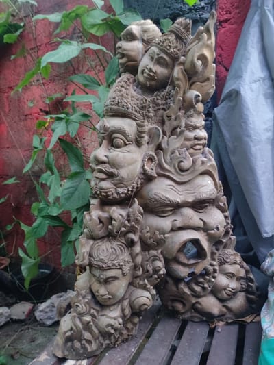 The image depicts a weathered, multi-faced stone sculpture, likely a representation of deities or mythological figures, sitting outdoors in what appears to be a garden or yard. The sculpture is composed of several stacked faces, each with distinct expressions and ornate detailing. The stone is beige or light brown, suggesting age and exposure to the elements. Behind the sculpture, there is a red brick wall partially covered with green foliage. To the right, a gray tarp is visible. The location is indicated as Bedulu, Indonesia, suggesting the sculpture is of Balinese or Indonesian origin. The sculpture is sitting on a wooden, slatted surface.