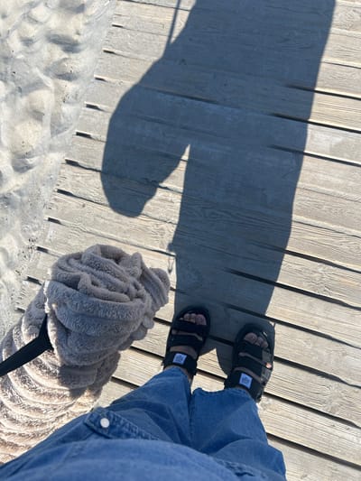 The image shows a person's feet wearing black sandals with white tags, standing on a wooden boardwalk. The person is wearing blue jeans. In the foreground, a fluffy, light-colored object, possibly a dog or a dog accessory like a blanket, is resting on the boardwalk. The boardwalk is situated next to a sandy area, suggesting a beach or coastal location. A long shadow is cast across the boardwalk, indicating it is daytime, likely late morning or afternoon given the angle of the sun. The weather appears to be sunny and bright. The scene is casual and relaxed. No visible text is clearly legible on the objects.