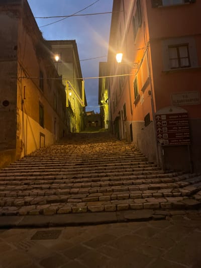 The image shows a set of stone stairs in Portoferraio, Italy, at the coordinates 42.816, 10.331. The stairs lead up a narrow street lined with old buildings on either side. The buildings are lit by streetlights, casting a warm glow on the stone steps.  The street is empty, and the only indication of life is a street sign on the right side of the image, pointing to various attractions in the town. The street sign, "Via Elbano Gasperi," is attached to a yellow building.  The "N" is a string of yellow lights, strung across the street. The sky is dark, but illuminated by the lights of the streetlamps and buildings. The scene is quiet and peaceful, giving a sense of solitude.