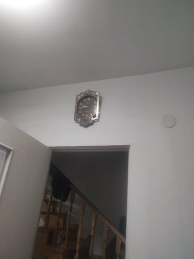 This media file shows an indoor scene, likely a home or hallway in Qaskeleñ, Kazakhstan. The primary object in focus is an ornate, vintage-style clock mounted on a white wall. The clock has a decorative, possibly metallic frame and a dark face with Roman numerals. The hands of the clock appear to indicate a time slightly after 4:00. To the left of the clock, a staircase with wooden railings and steps is visible, leading downwards into a dimly lit area. A dark garment, possibly a coat or jacket, is draped over the railing of the staircase. On the wall to the right of the clock, there is a circular, white object, possibly a light fixture or a decorative element. The lighting in the scene is dim, suggesting it might be evening or a poorly lit interior. There is no visible text in the image.