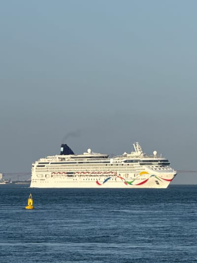 A large white cruise ship, the Norwegian Dawn, sails on a calm, deep blue ocean under a clear, pale blue sky. The ship features a distinctive rainbow design along its hull and the text "NORWEGIAN DAWN" is visible. A black plume of smoke emerges from the ship's funnel. In the foreground, a yellow buoy with a black top floats on the water. In the distance, a suspension bridge and the faint outline of a city, likely Oeiras, Portugal, are visible on the horizon. The overall scene depicts a serene day at sea.
