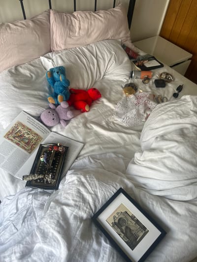 The image depicts a cluttered bedroom scene, specifically a bed with white linen duvet cover and two light pink pillows against a black metal headboard. To the right, a wooden wall and a white nightstand are visible.
On the bed, a blue plush teddy bear is seated with its hands near its face, beside a red plush teddy bear lying on its side, and a lavender plush teddy bear also lying on its side. An open magazine or journal titled "COLONIAL AND ANTI-BLACK LEGACIES OF FRAGRANCE AND DEODORIZATION" lies face-up, displaying text and an ornate illustration. A second open magazine is partially visible beneath it.
Resting on the open magazine is a black rectangular tray with gold decorative trim, holding various small items: a white skull charm, a purple butterfly ornament, a miniature metallic violin, a bundle of white sage, and a silver charm resembling an intricate animal.
Further to the right, towards the nightstand, are several personal items scattered on the bed and nightstand: a silver and orange can (possibly a beverage), a gold and clear glass perfume bottle, a small black box, a tray containing rings and other jewelry, a black electronic device, and a white lace garment with red accents underneath.
At the bottom right of the bed, a black-framed picture with a white mat displays a monochrome image of a cathedral facade, identifiable as "TARRAGONA Catedrale" from faint text on the original postcard within the frame.
The precise location within the city of Wealden, United Kingdom, cannot be determined from the visual information provided in the image.
