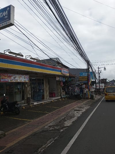This image depicts an urban street scene in Vaatharawattai, Sri Lanka, under an overcast sky, viewed from a slightly elevated perspective on the left side of a paved road. On the left, a single-story commercial building dominates the foreground, featuring a distinct red, blue, and yellow striped awning and facade. This building is identified by a prominent "Indomaret" sign and a partially visible "ATM+" sign with "ATM Bersama" text. Its storefront consists of large glass panels displaying various products and promotional posters. A dark-colored scooter is parked in a designated yellow-marked parking space directly in front of the entrance, near a cylindrical trash bin.
Adjacent to the Indomaret, a smaller vendor stall displays an assortment of goods, including balloons and packaged items. Another motorcycle is parked near this stall. Further down the street, a utility pole heavily laden with a dense bundle of black electrical and communication wires rises on the left, with additional wiring extending across the street. A blue sign advertising "AYAM GORENG MBAK SINTA" is visible further along the left side. The asphalt road features a solid white line on its right edge. In the distance, a yellow minivan or taxi is visible on the road, traveling away from the viewer. No individuals are clearly discernible within the frame.
