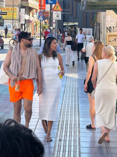 The image depicts a street scene in Chania, Greece. In the foreground, a man wearing bright orange shorts and a towel draped over his shoulders is walking next to a woman in a white dress. The man is holding a bottle of beverage, and the woman is also holding a bottle. They appear to be walking on a tiled sidewalk with a tactile paving strip. In the background, other pedestrians are visible, along with buildings, street signs, and a bus. The atmosphere suggests a sunny day, possibly during tourist season, given the casual attire of the people. The architecture and signage hint at a Mediterranean setting.