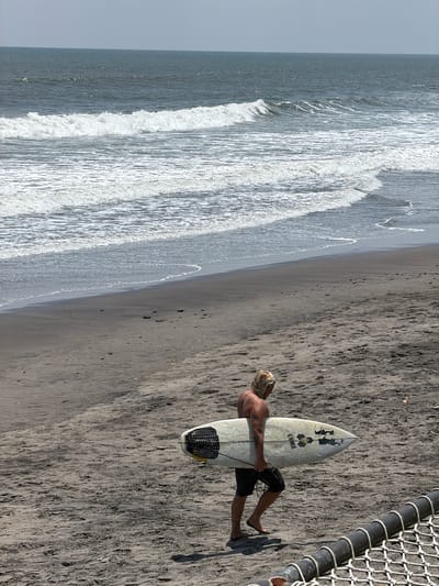 A male individual with blonde hair, wearing dark shorts, walks barefoot across a dark sand beach in Belalang, Indonesia. He carries a white surfboard, featuring a black traction pad and "Al Merrick" branding, under his right arm. Tattoos are visible on his left arm. The background depicts the ocean with multiple white-capped waves breaking towards the shore, under a pale grey sky. The dark sandy foreground contains scattered pebbles and visible footprints. A dark cylindrical barrier with white netting is partially visible in the lower right corner.