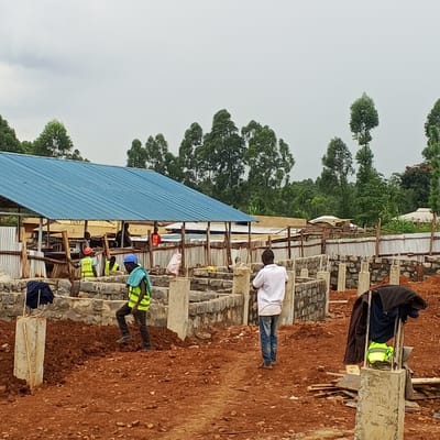 This media file captures an active construction site in Borabu Chitago ward, Kenya, under an overcast sky. The ground is predominantly exposed reddish-brown soil, characteristic of the region, indicating ongoing earthworks.
In the foreground and mid-ground, the foundations of a new building or complex are visible. These foundations consist of rough-hewn stone blocks mortared together, forming low walls, interspersed with concrete pillars. Several workers are present on the site. On the left foreground, a worker wearing a blue hard hat, a yellow safety vest, and dark trousers walks across a mound of dirt. In the mid-ground to the right, another person in a white t-shirt and blue jeans walks away from the camera.
Towards the background, a large structure with a bright blue corrugated metal roof stands, possibly a temporary shelter, storage area, or an existing building being expanded. Beneath this roof, several other workers are visible, some also wearing yellow safety vests. Corrugated metal sheets are used as fencing or temporary partitions around parts of the site. In the distance, a dense line of green trees suggests a verdant, possibly rural or semi-rural, setting.
Notable details include construction materials like wooden planks scattered on the ground. A dark jacket, a yellow safety vest, and a blue towel-like item are hanging on one of the concrete pillars in the right foreground. Faint, partially obscured text, possibly "TENIS" or "TENTG," can be seen on a banner or sign attached to the blue-roofed structure in the background. The scene depicts a busy construction environment in broad daylight.