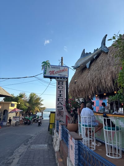Here is a description of the image:

The image depicts the Motel Mexicola in Canggu, Indonesia. The motel features a thatched roof with a decorative fish mounted on it. Patrons are seated in white chairs on a patio, with a bar visible in the background. A sign reading "Motel Mexicola" is displayed prominently. Below it, a sign reading "Boticas" is affixed. The street scene shows motorcycles and pedestrians. The ocean is visible in the distance. The sky is blue.