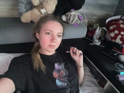 A young Caucasian woman with light brown hair pulled back in a ponytail and fair skin is lying down on a bed, taking a selfie. She is wearing a black t-shirt with a colorful abstract graphic on the front. In her right hand, she is holding a slender, dark-colored electronic device. The background includes several stuffed animals, a bouquet of red and white artificial roses, and various personal items on a black surface, possibly a bedside table. The lighting suggests an indoor setting, likely a bedroom. The woman appears to be in a relaxed posture. The image does not provide any specific details to place it in Makiivka, Ukraine.