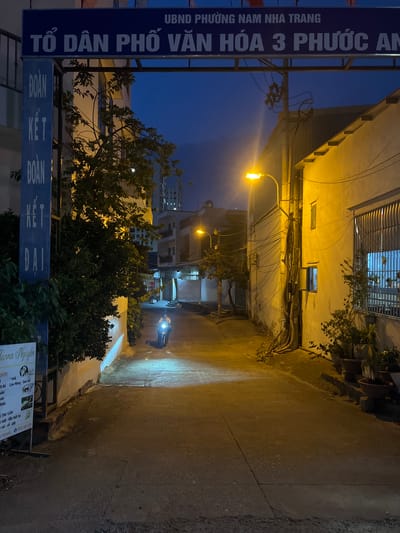 The image shows a narrow alleyway in Nam Nha Trang, Vietnam, at dusk. A person on a motorbike approaches from the distance. Streetlights illuminate the scene, highlighting the concrete walls and ground of the alley. A sign with the inscription "TỔ DÂN PHỐ VĂN HÓA 3 PHƯỚC AN" is visible, indicating the neighborhood. Wires crisscross overhead. A smaller sign, labeled "Larna Nguyễn," appears on the right foreground, advertising nail and spa services. Trees and plants are visible on both sides of the alleyway.