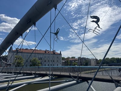 The image shows a view from the Bernatka Footbridge in Kraków, Poland, overlooking the Vistula River. The bridge, a modern structure with a distinctive curved design, is made of steel and has a metal railing. The image is taken from the bridge, looking toward the river, with a white building with red roof tiles visible in the background. The bridge is empty except for two figures that appear to be sculptures of people performing acrobatics. Both figures are suspended in mid-air, one on a chair and the other holding onto a cable. The background features a blue sky with white clouds, and a river. The sky is clear with no visible pollution. The river water is calm, and the riverbank is lined with trees.