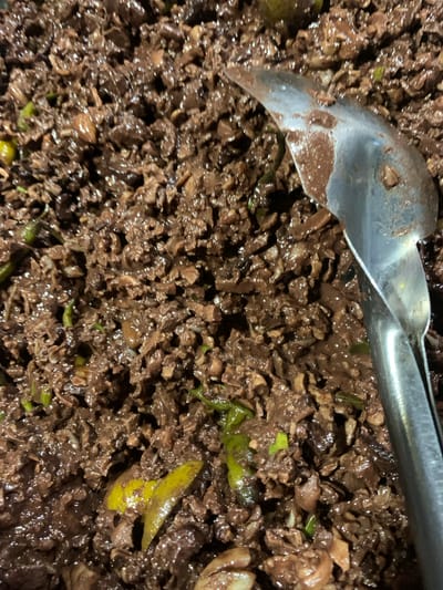 This image shows a close-up, overhead view of a dark, finely chopped dish being served with a metal spoon in Kiblawan, Philippines. The dish appears to be a savory mixture, possibly meat or blood-based, with visible small green pieces, likely chili or herbs, and some yellow slivers, possibly ginger or lime. The texture is moist and slightly clumpy, with a glossy sheen. The spoon, made of polished metal, is positioned in the upper right quadrant of the image, with its edge resting in the food, suggesting it is in the process of being served or is already in use. The lighting is dim, creating shadows and highlighting the textures of the food. The overall impression is of a hearty, possibly traditional, dish being prepared or consumed in a domestic or communal setting in Kiblawan.
