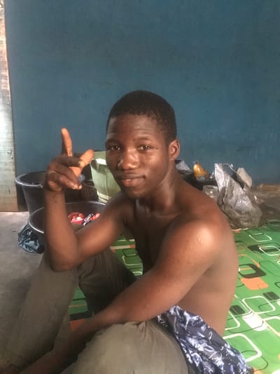 A shirtless young man with dark skin sits in front of a green-patterned wall and a blue wall. He wears grey pants and a blue patterned cloth around his waist. He is smiling and holding his hand out with his index finger extended. Behind him are some plastic containers and other objects covered with a grey plastic bag. The location appears to be an indoor space. The city of Ilorin, Nigeria is not discernible from this image.