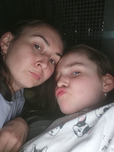 Two individuals, likely a mother and daughter, are positioned close together and looking at the camera. The younger individual is resting their head on the older individual. The younger individual is partially covered by a white blanket with cartoon dogs printed on it. The older individual is wearing a light blue shirt. Their facial expressions are similar, both with pursed lips. The background shows a wall with a grid pattern on the left side. The location is Krasnogorskoye, Russia.