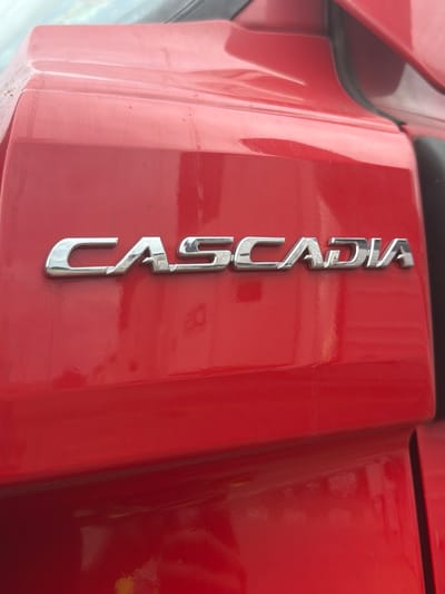 A red vehicle's exterior features the chrome lettering "CASCADIA" on its side. The letters are metallic and reflective, with the word "CASCADIA" clearly legible. The vehicle's surface appears to be clean and well-maintained, with a glossy finish. The background is out of focus, but suggests an outdoor environment, possibly a parking lot or roadside. No people or other objects are discernible in the image. The scene is static and does not depict any action. The location in Murfreesboro, United States of America, is not visually indicated within the image itself.