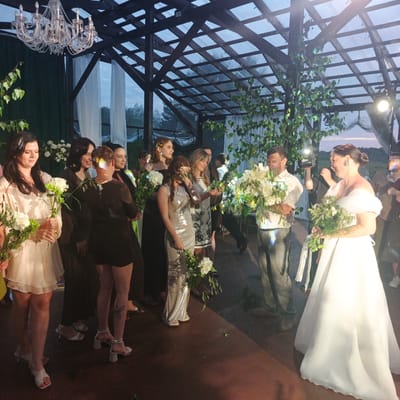 The image depicts a wedding or special event held outdoors under a covered wooden structure with a transparent roof. The scene appears to be at dusk, with artificial lighting and natural light from the sky visible.
In the foreground, a bride in a white off-the-shoulder wedding dress is the central figure. She is holding a bouquet of white flowers and appears to be interacting with someone off-camera to her right. To her left stands a man in a light-colored shirt and grey pants, also holding a large bouquet of white flowers. He is looking towards the bride and smiling.
To the left of the bride, several women are gathered. One woman in a cream-colored dress is holding a bouquet. Another woman in a dark dress is laughing. Further left, another woman in a sequined dress is holding a bouquet.
The environment includes a dark wooden structure with visible beams and transparent panels in the roof. Green foliage and white drapery are visible in the background. A large crystal chandelier hangs from the structure. The ground appears to be a dark, reddish-brown surface. The overall atmosphere suggests a festive gathering.