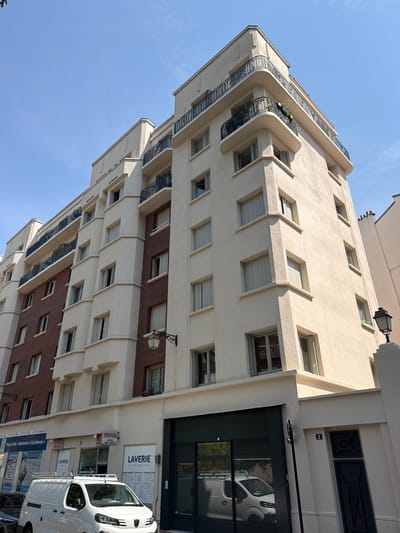 This is a vertical, eye-level photograph of a building facade in Courbevoie, France, on a clear, sunny day. The building is a multi-story structure with a light-colored, textured exterior. The architecture features rounded balconies with black railings on the upper floors, and a series of recessed windows with angled, cream-colored frames on the lower floors. Below these, a section of the building is clad in red brick, also with windows.

The ground floor appears to be commercial, with a dark-colored entrance door reflecting a white van. Several signs are visible: one prominently displays "LAVERIE" (laundry) with text in French indicating rapid cycles and quick drying. Another sign reads "MACHINES A LAVER" (washing machines). Further down, a sign for "Point d'Info Stationner à Courbevoie" (Information point, Parking in Courbevoie) is visible, along with parking-related symbols. A white van is parked in front of the building, partially obscuring the ground floor. A black lantern-style light fixture is attached to the building near the top. The bright blue sky suggests it is daytime. The overall impression is of an urban street scene in a residential and commercial area.