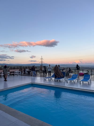 A rooftop pool area in Mukhatskaro, Georgia, is depicted at dusk, with a group of people socializing and dining. The foreground features a bright blue swimming pool with a tiled deck surrounding it. Beyond the pool, several lounge chairs are arranged on the deck. In the middle ground, a group of individuals are gathered around tables and seating areas, some standing and some seated. A long structure with red and white striped seating is visible. In the background, a power line tower stands tall against a sky filled with soft pink and blue clouds. The landscape beyond appears to be rolling hills and a distant town. The overall atmosphere suggests a social gathering or event taking place at a scenic overlook.