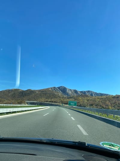 The image displays a multi-lane asphalt highway on a clear, sunny day, viewed from the interior of a vehicle traveling towards Pelev Brijeg, Montenegro. The sky, a deep clear blue, occupies the upper half of the frame, with a prominent vertical light streak or reflection visible on the left side of the windshield.
In the mid-ground, a range of mountains stretches across the horizon. The mountains on the left are covered with vegetation exhibiting autumnal or dry tones, while the mountains on the right feature more exposed, rugged grey rock faces.
The highway comprises at least two lanes in the direction of travel, marked by white dashed lines. On the left, a white metal guardrail follows the curve of the road, and a concrete overpass or bridge structure is visible in the distance. On the right, another white metal guardrail borders the road, beyond which is a vegetated slope leading to the mountains.
A green overhead road sign is positioned on the right side of the highway. It indicates "Montenegro Pelev Brijeg" in Latin script, along with Cyrillic text, symbols for a gas station and restaurant, and the distance "2.4 KM".
The foreground shows the black dashboard and the lower edge of the vehicle's windshield. A windshield wiper blade is partially visible, and a circular green and white sticker is affixed to the lower right portion of the windshield. No individuals are visible in the scene.