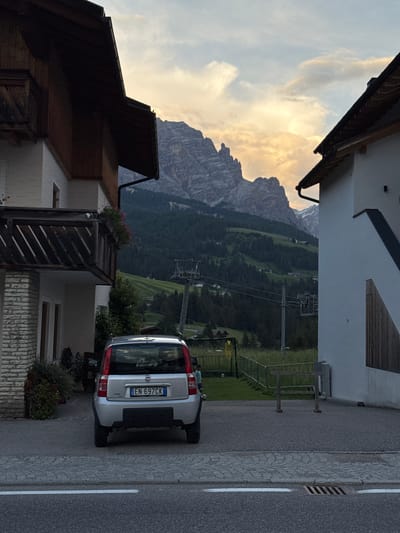 In the foreground of the image, a silver Fiat Panda car is parked on a paved surface. The car's license plate is visible and reads "EN 697CK". To the left of the car, a portion of a building is visible, featuring white brickwork and a wooden balcony. To the right, another building with white walls and dark wooden siding is partially visible. The background displays a dramatic mountain landscape with lush green hillsides dotted with trees and some scattered buildings. A ski lift structure is also present on the hillside. The sky is filled with clouds, illuminated by a soft, warm light, suggesting either sunrise or sunset. A small part of the street with a white line marking and a drain grate is visible at the bottom of the image. A child is visible in the far right of the image. The scene appears to be a residential area in the Italian Dolomites.