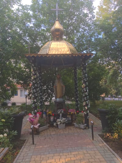 This image depicts an outdoor shrine, likely in the city of Zalishchyky, Ukraine. The shrine features a golden dome topped with a cross, supported by dark pillars adorned with white artificial flowers. At the center of the shrine stands a statue of a woman in a beige robe, believed to be the Virgin Mary. Surrounding the shrine are various floral arrangements in planters and on the ground, including bouquets of pink, white, and red flowers, as well as potted plants. The pathway leading to the shrine is paved with light-colored bricks. In the background, trees with lush green foliage frame the scene, and a building with a light-colored facade and a red roof is partially visible. The overall environment suggests a place of quiet contemplation and religious significance within a residential or public urban space.