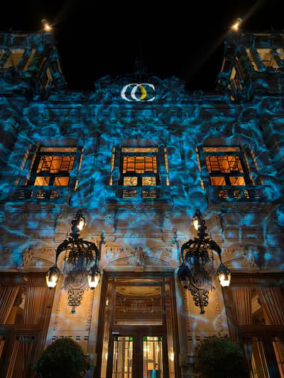 The image shows the exterior of the historic Casino Municipal in San Pellegrino Terme, Italy, at night. The building's facade is illuminated with a vibrant blue and green light projection, creating a dynamic, wavy effect across the stonework. The ornate architecture of the building, with its detailed carvings and decorative elements, is visible beneath the projection. Three large windows with dark, intricate frames are prominent on the upper level, and their interiors are warmly lit with an orange glow, suggesting activity within. Below these, decorative wrought-iron balconies and two large, ornate lamps with glowing bulbs are mounted on the facade. The lamps are suspended from elaborate metal brackets. The entrance to the building is centered, with large wooden doors and adjacent glass panels. Above the entrance, a sign reads "CASINO MUNICIPALE S. PELLEGRINO T.". To the left and right of the entrance, tall windows are covered by heavy, textured curtains. A dark green bush is positioned in the foreground, partially obscuring the lower section of the building. The sky above is dark, with a single bright light source emanating from the top of the building, casting a beam upwards. A white and yellow logo, possibly an emblem or artistic representation, is projected onto the very top of the building's structure.
