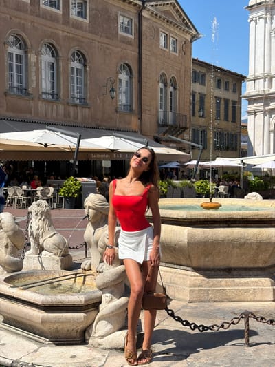 A woman poses in front of a fountain in Mozzo, Italy. The woman is wearing a red tank top and a white skirt. She is holding a brown purse and wearing brown sandals. The fountain is made of stone and has statues of lions. In the background, there are buildings with shops and outdoor seating. The sky is blue and there are some clouds. The woman is smiling and looking at the camera. The scene is brightly lit by the sun.