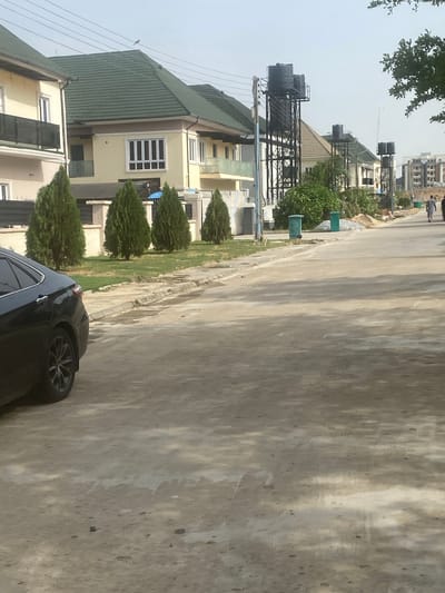 The image depicts a street in Iddo Tudunwada, Nigeria. The scene is dominated by a paved road occupying the majority of the frame. A black car is partially visible in the lower right corner. Houses are seen along the left side of the street, featuring green roofs and light-colored walls. Cylindrical trees are planted in front of the houses. A water tower is visible behind one of the houses. In the background, pedestrians are seen walking along the road.