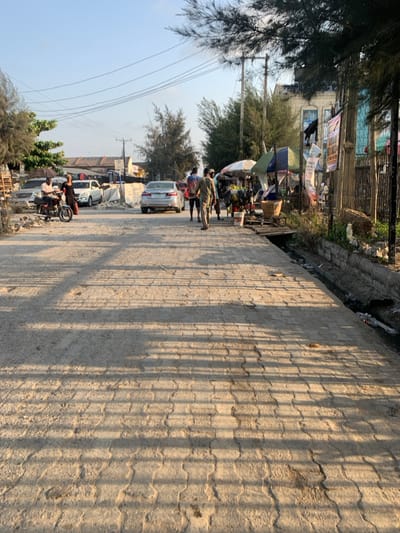 The image depicts a street scene in Ikota, Nigeria, under clear daylight, casting long shadows indicative of morning or late afternoon. The foreground features a wide, paved road constructed of interlocking concrete blocks, showing signs of dirt and wear.
On the left side of the road, a person in a light-colored shirt is seated on a brown motorcycle. Nearby, another individual in a dark dress stands on the paved surface. Behind them, multiple vehicles are visible, including a silver SUV and a white sedan, parked amidst piles of wooden pallets and construction debris. Utility poles with overhead power lines are present on this side.
In the center of the road, a silver sedan is either parked or moving slowly away from the camera. A group of three individuals walks away from the camera in the middle-right of the paved area; one wears a striped shirt, another a patterned garment.
The right side of the street features a series of makeshift market stalls operating under two distinct, colorful umbrellas (one white and green, one blue and white). Various goods, including baskets and produce, are displayed. A narrow, concrete-lined drainage ditch runs parallel to the road, containing dark liquid and refuse. A rudimentary wooden plank bridge crosses this ditch to access one of the stalls. Behind the stalls, a tall fence constructed of bamboo or similar reeds is visible, adorned with several banners or posters. A multi-story building with light-colored walls and blue-framed windows stands further back. Tall, slender trees with sparse foliage line both sides of the street, and additional utility poles with power lines are present on the right.