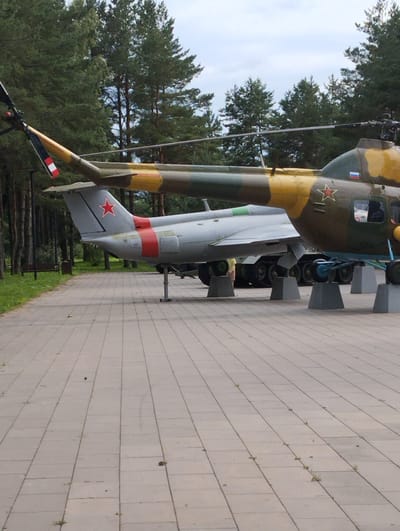 The image shows two military aircraft on display, likely in a museum or memorial park, in Kingisepp, Russia. The primary subject is a helicopter, painted in a camouflage pattern of green, yellow, and brown. It has a red star insignia on its side and a Russian flag decal on its tail. The helicopter is supported by concrete blocks. Partially visible in front of the helicopter is a jet aircraft, painted in a light gray color with a red star insignia on its tail and red stripes along its fuselage. It also appears to be on display.

The scene is set outdoors, with a paved walkway in the foreground composed of rectangular concrete tiles. The background consists of a dense forest of tall, slender pine trees, suggesting a temperate climate and possibly daytime given the ambient light. The sky is overcast with clouds, indicating possibly cool or mild weather. There is a wooden bench and a trash receptacle visible near the edge of the treeline on the left side of the image, further suggesting a public park or exhibition area.

No people are directly visible in the image, though a person is partially visible near the jet aircraft in the lower left portion of the image, wearing a light yellow top. The overall mood is one of historical preservation and perhaps remembrance. There is no visible text in the image other than the insignias on the aircraft.