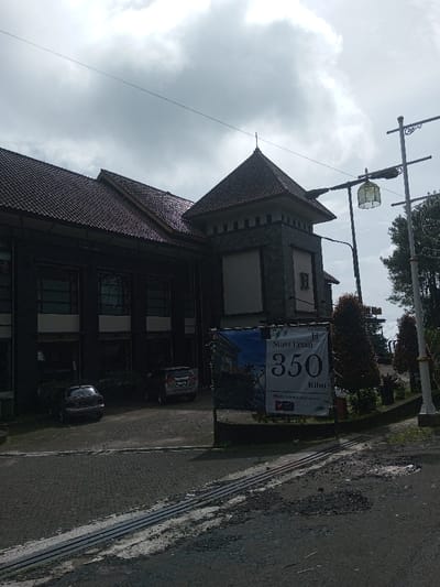 This image captures an outdoor scene in the city of Sandamal Eliya, Sri Lanka, under an overcast sky during daylight. The primary subject is a multi-story building with a dark facade, possibly dark grey or brown, and a dark tiled, pitched roof. A distinctive tower-like section on the right side of the building features a pyramidal tiled roof, a stone-textured exterior, a large white rectangular panel, and a dark 'H' symbol.
In the mid-ground, two vehicles are parked on a paved area in front of the building: a dark-colored sedan on the left and a silver SUV slightly to its right. The foreground features a large vertical banner on a stand, displaying "Start From 350 Ribu," "propertyindahabadi.com," and a partial logo, against a blue and white background. To the right of the banner, landscaped areas with green and reddish-brown foliage are visible. An ornate streetlight with a lantern-style fixture is positioned behind the banner, alongside a plainer street light pole. The ground consists of a paved area transitioning to a darker asphalt road in the immediate foreground, showing signs of wear and tear and featuring embedded parallel strips, possibly drainage or tracks. No individuals are visible within the scene, and no discernible actions or interactions are occurring.