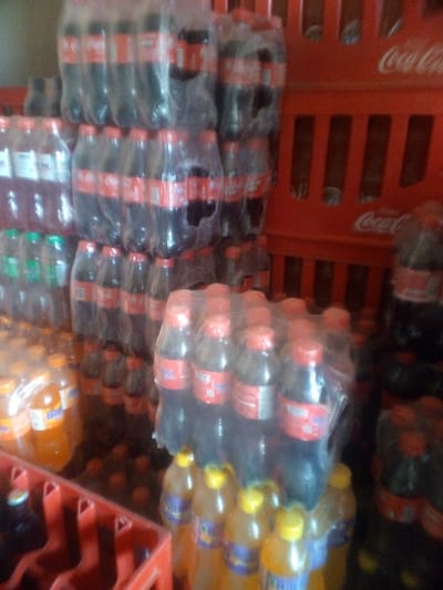 The image displays a densely packed storage area, likely a backroom or a section of a beverage retail establishment. The primary subjects are numerous plastic bottles of various soft drinks, stacked and arranged in bundles and crates.
The scene is dominated by large quantities of Coca-Cola bottles, identifiable by their dark liquid, red labels, and red caps. These bottles are bundled together in clear plastic wrap and stacked high, forming significant columns of inventory. The iconic "Coca-Cola" logo is also clearly visible on several red plastic crates positioned on the right side of the image.
Interspersed with the Coca-Cola products are other brands of soft drinks. Bundles of bottles containing reddish and green liquids are visible, as are several bundles of orange or yellow-colored beverages. Some of these orange/yellow drinks explicitly show the "Bigi" brand name on their labels and feature yellow caps.
The drinks are stored in a functional manner, with bundles stacked directly on top of each other and some bottles placed within red plastic crates. The crates themselves appear sturdy and are typical for beverage transport. The lighting is ambient, suggesting an indoor setting without direct sunlight. There are no people visible, indicating a snapshot of a static inventory rather than an active transaction or stocking process. The overall impression is one of a well-stocked beverage supply point.