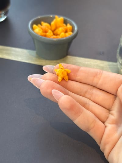 In Durbuy, Belgium, a person's hand, adorned with nude-colored nail polish and manicured nails, holds a single, small, star-shaped, orange-colored crunchy snack. In the background, a dark gray bowl overflows with similar snacks. The snacks are on a light-colored wooden surface, which rests on a dark gray tabletop. The scene suggests a casual snack break.